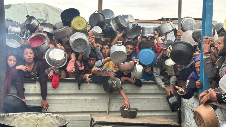 Children wait in line for food with pots in hand.