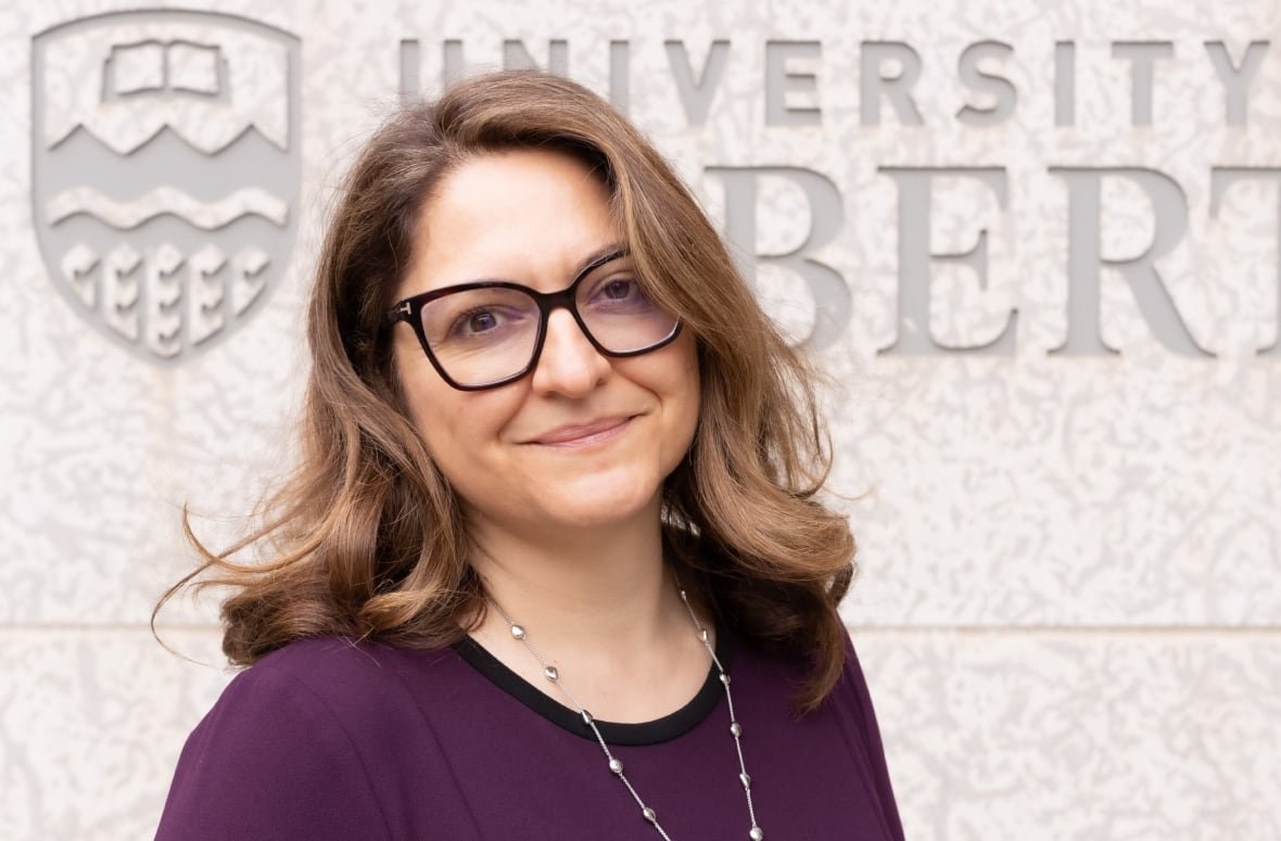 A woman poses in front of a wall where the name University of Alberta is engraved.