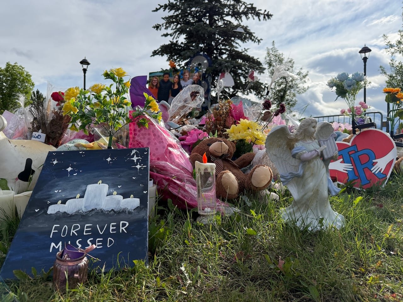 outdoor memorial in front of a spruce tree with flowers, candles, teddy bears, photos of children, and an angel statue beside a sign that reads “FOREVER MISSED”