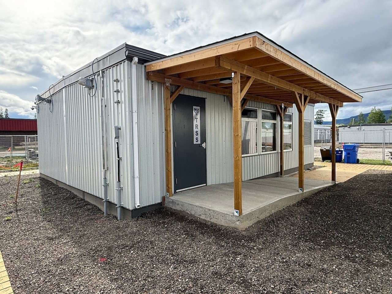 small silver corrugated-metal portable building on a gravel lot with a wooden porch, windows, and a sign on a black door door that reads "TRSS"