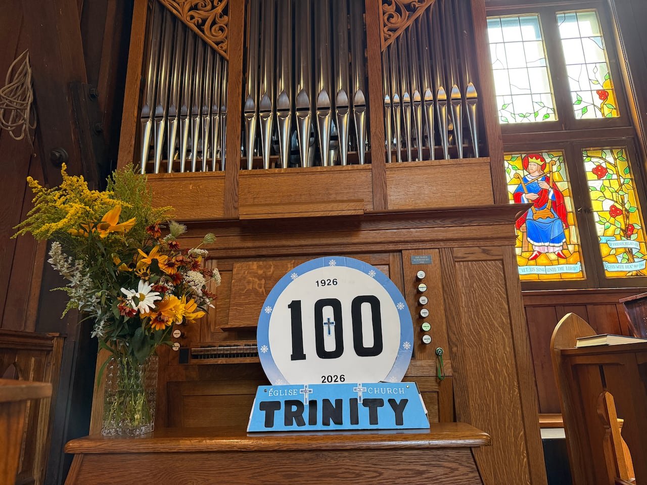 A cardboard sign marking 100 years sits on the bench of a church organ with the metal pipes visible behind the sign.