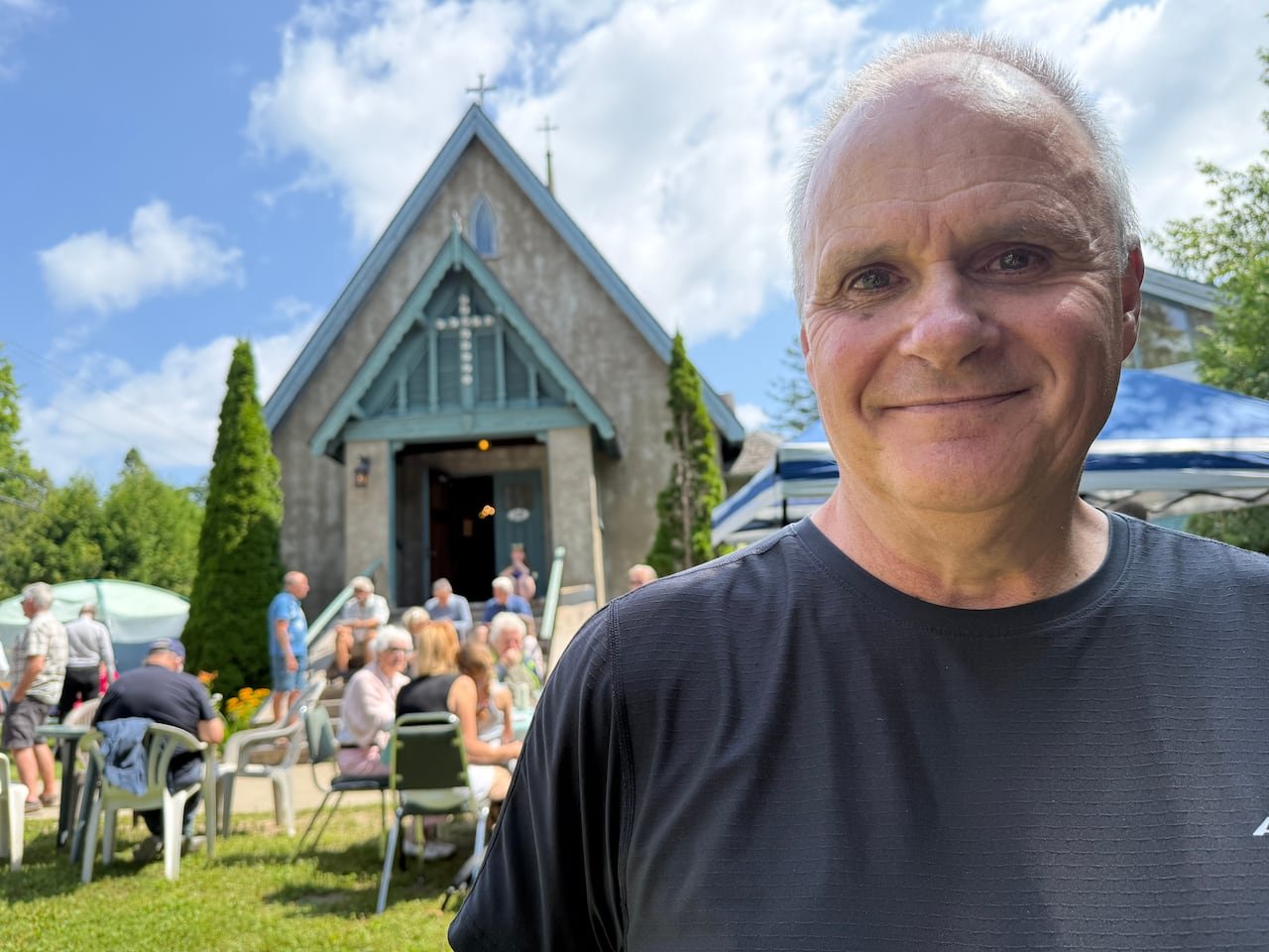 white man with blue eyes and blue shirt looks at the camera with an old church in the background and people milling about in front of the church